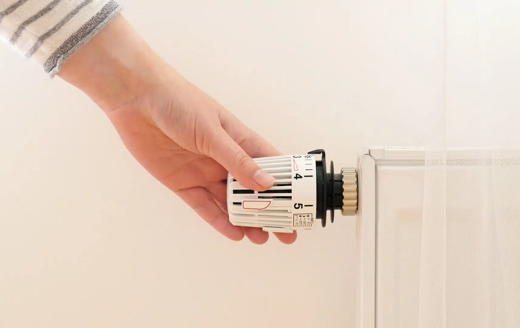 Effiziente Energienutzung: Hand reguliert die Heizungstemperatur Eine Hand dreht am Thermostat einer Heizung, um die Temperatur einzustellen. Das Bild symbolisiert Energieeffizienz und bewussten Umgang mit Ressourcen in einem minimalistischen, hellen Umfeld.