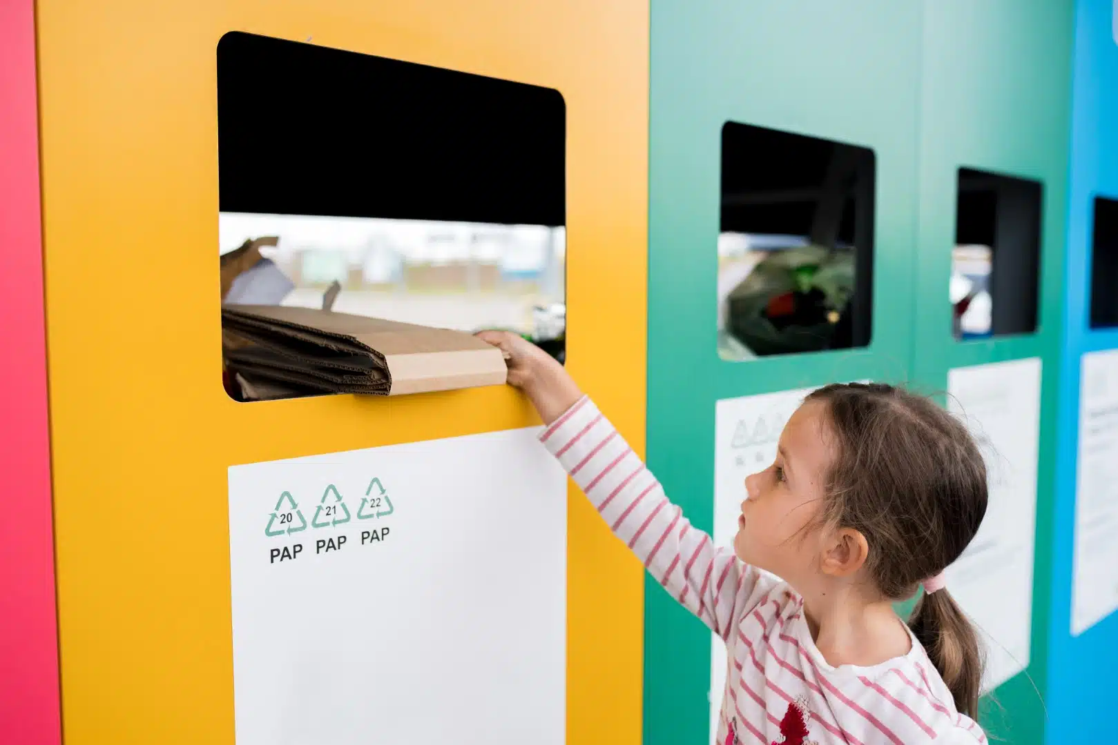 Mädchen recycelt Karton in der Papiertonne Kinder werfen Papier in die Recycling-Tonne Nachhaltiges Lebenskonzept Nationale Recyclingwoche Ein kleines Mädchen recycelt Karton, indem sie ihn in einen gelben Recyclingcontainer steckt. Die Container sind in verschiedenen Farben gestaltet und symbolisieren eine nachhaltige Mülltrennung. Das Bild zeigt umweltbewusstes Verhalten und den frühen Einstieg in Recycling.