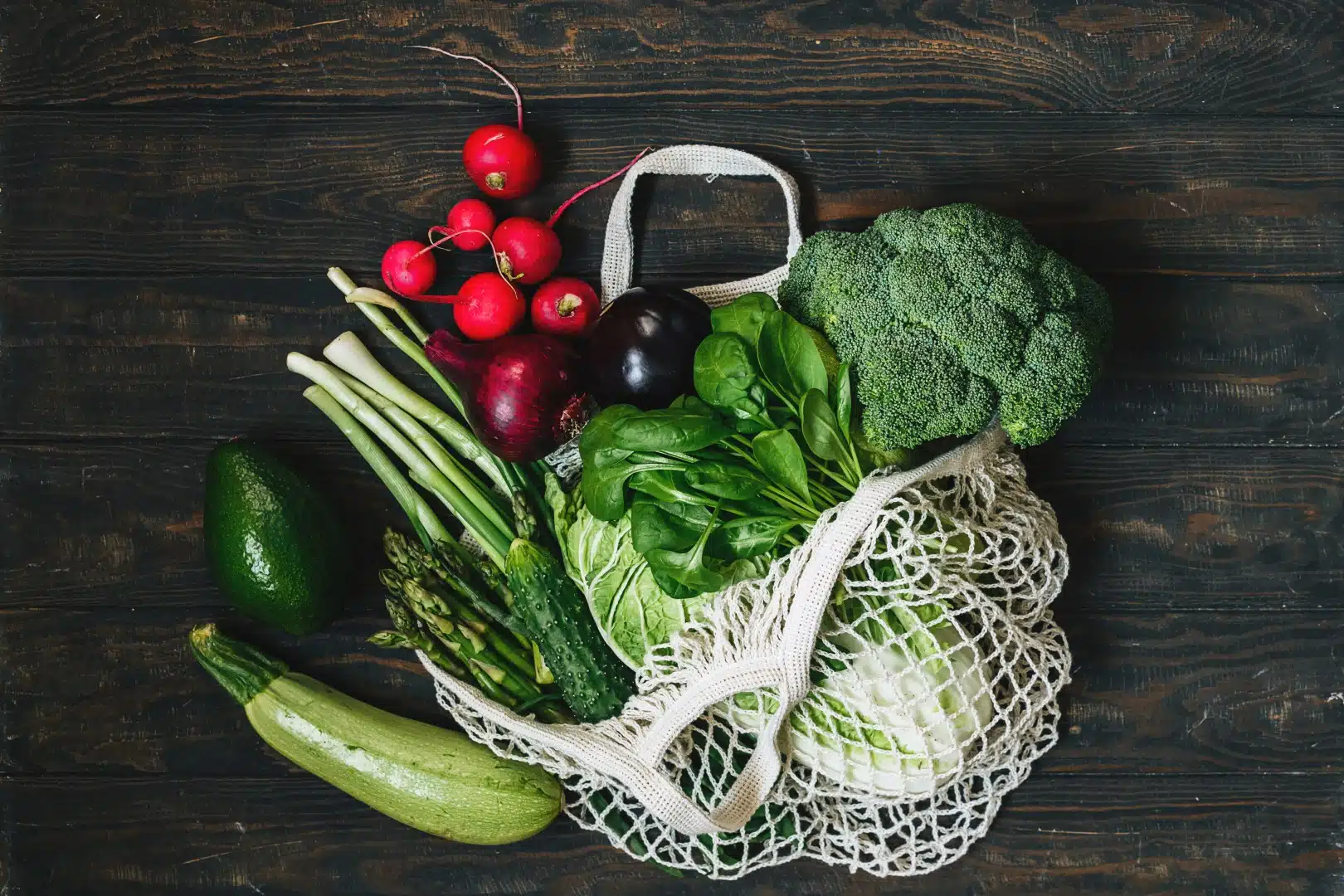Gemüse-Brokkoli-Salat Auberginen-Vogerlsalat in einer Netztasche Null Abfall gesundes Einkaufskonzept Das Bild zeigt frisches, farbenfrohes Gemüse, das in einem wiederverwendbaren Netzbeutel präsentiert wird. Auf einer dunklen Holzoberfläche liegen unter anderem Radieschen, Brokkoli, Avocado, Zucchini, grüner Spargel, Spinat und ein paar weitere grüne Gemüsesorten, die ein nachhaltiges und gesundes Einkaufen symbolisieren.