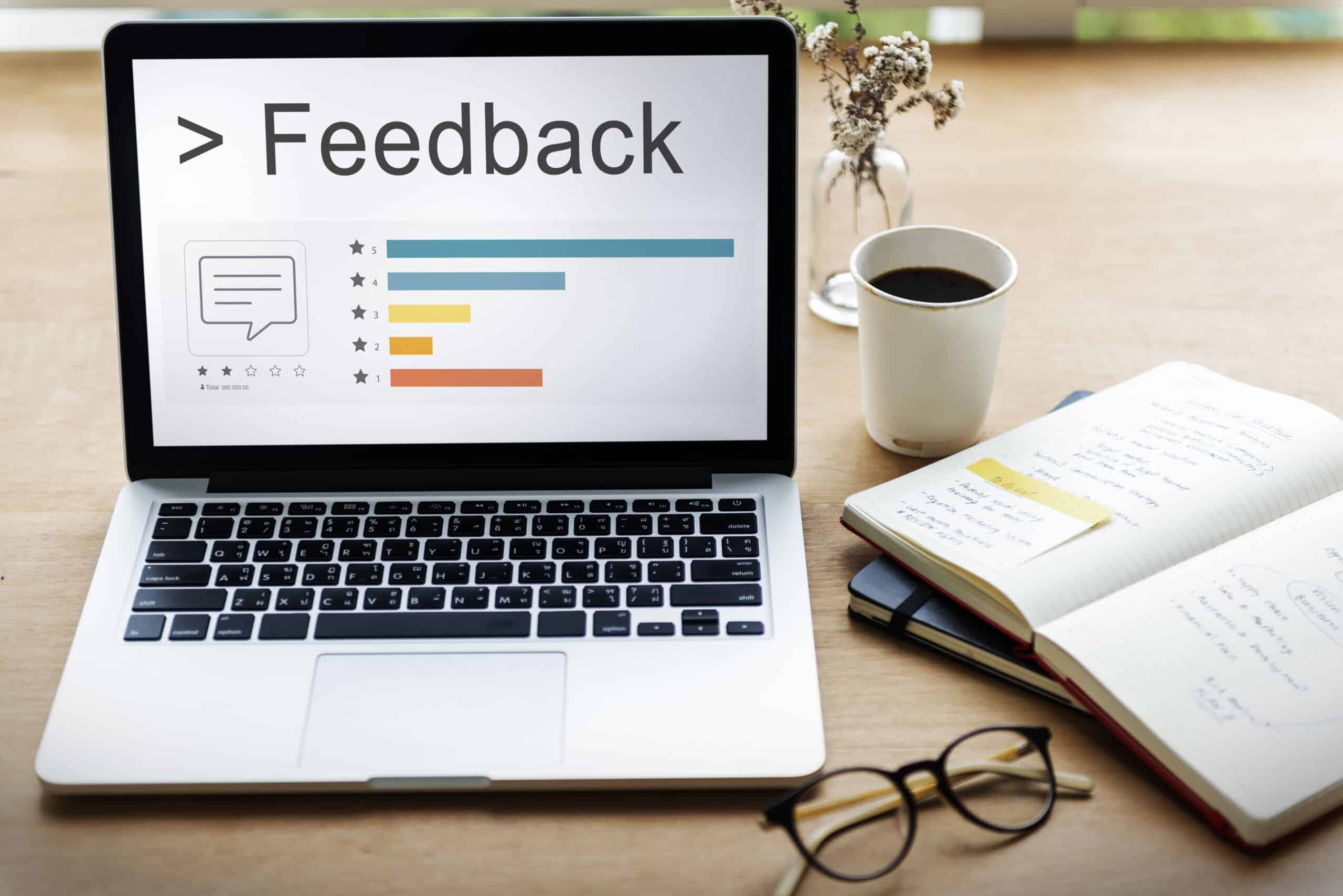 Laptop mit Feedback-Umfrage und Balkendiagrammen auf dem Bildschirm, daneben Notizbuch, Brille und Kaffeetasse auf dem Schreibtisch