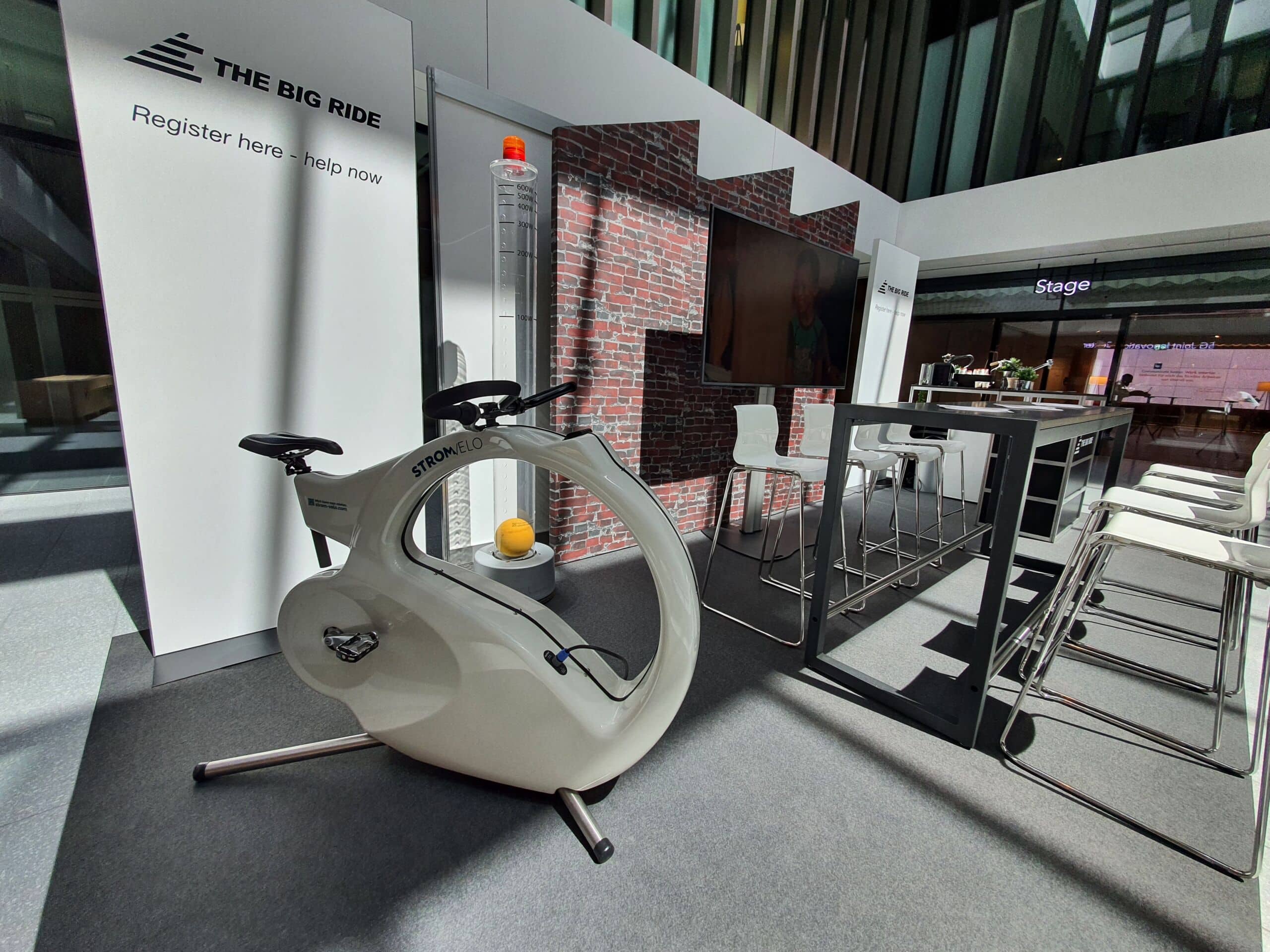 20220510_140655 | Strom-Velo Exhibition booth with a white stationary bike labeled Stromelo, brick-wall backdrop, large screen, and white seating area.