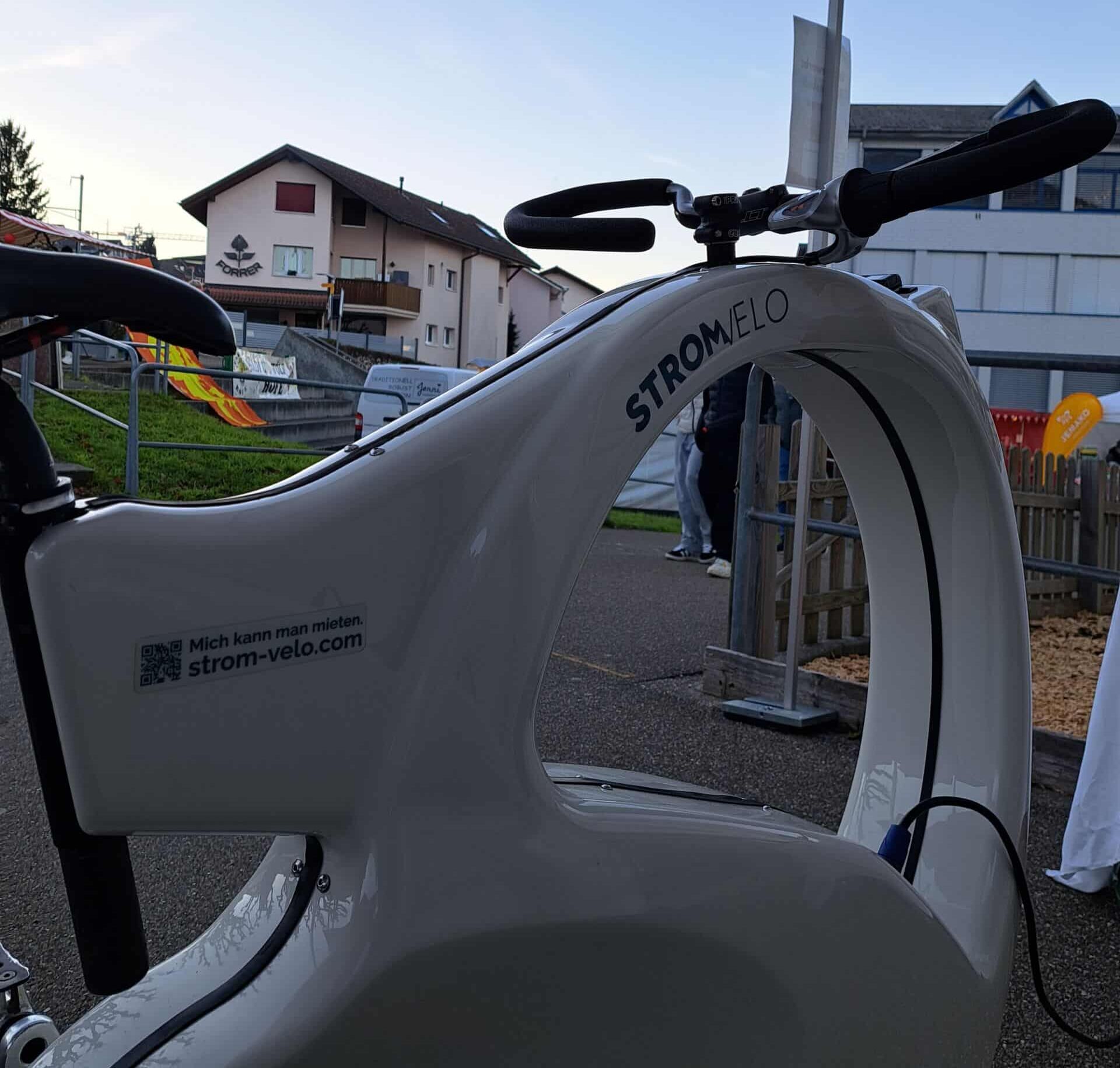 Close-up of a white streamlined Stromvelo bicycle at an outdoor booth, handlebars in view and a tent overhead; branding and a web URL visible on the bike.