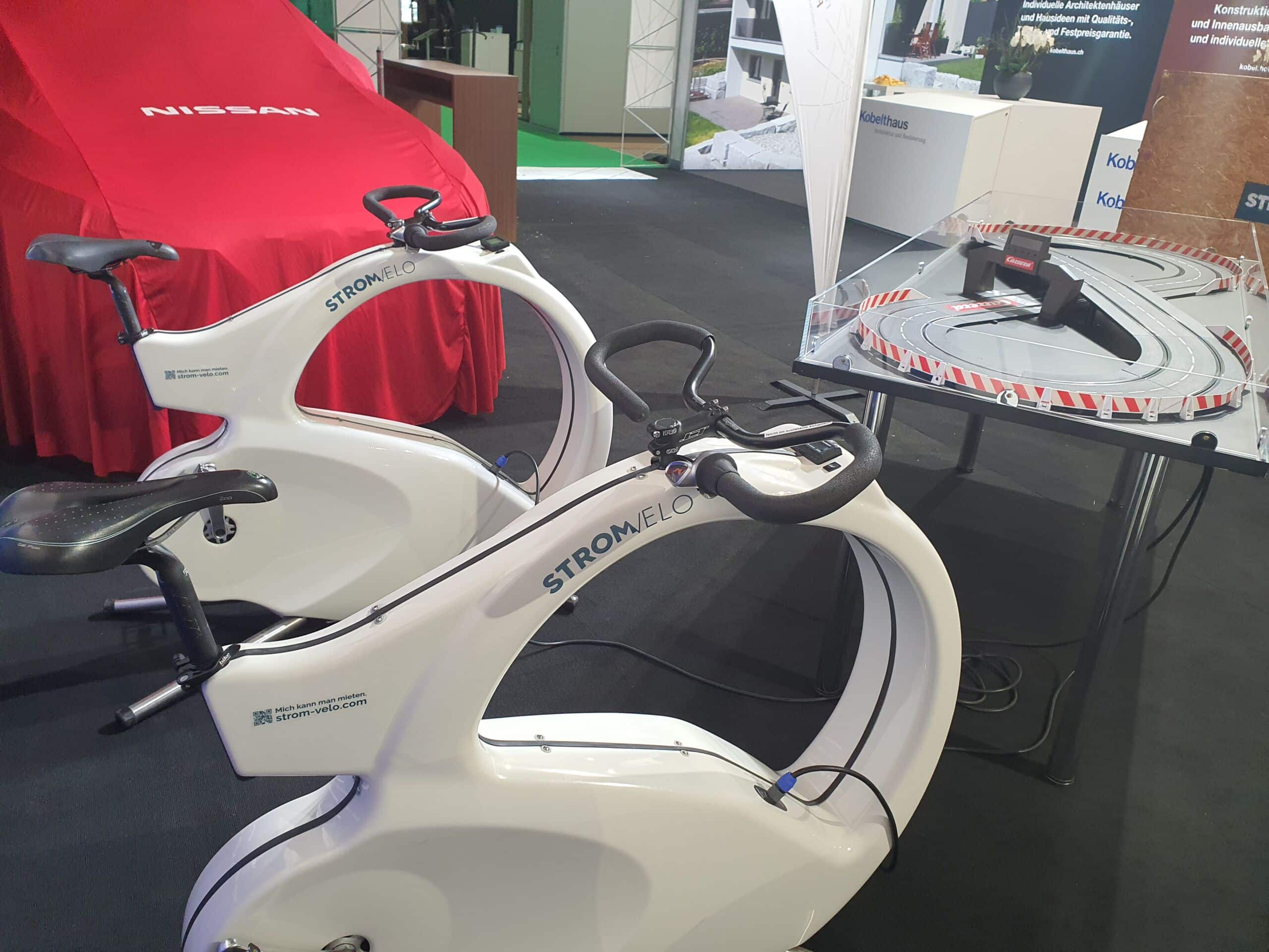 Exhibit booth with two white Stromvelo stationary bikes, a red Nissan-covered car in the background, and a circular track display behind.