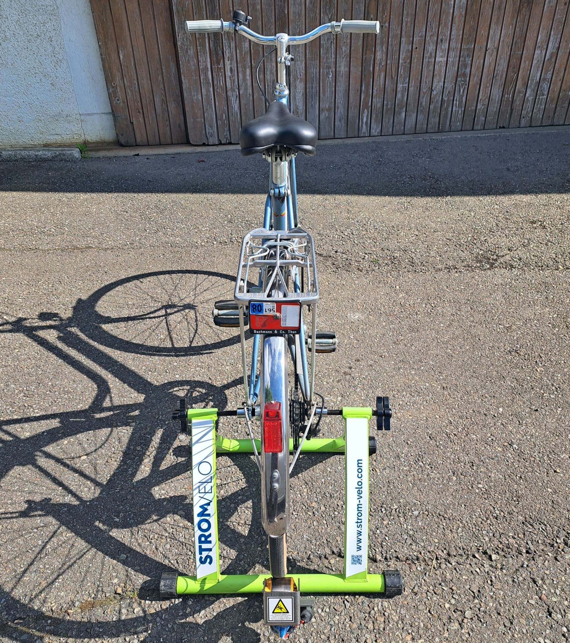 Top-down view of a blue bicycle on a bright green bicycle stand, with white panels and a rear light, parked on a gravel surface near a wooden fence.