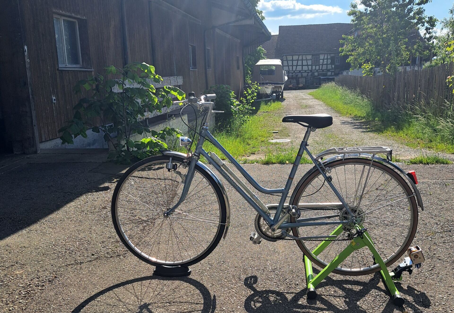 Two bicycles parked on a gravel path beside a dark wooden building on a sunny day, with shadows cast long on the ground.