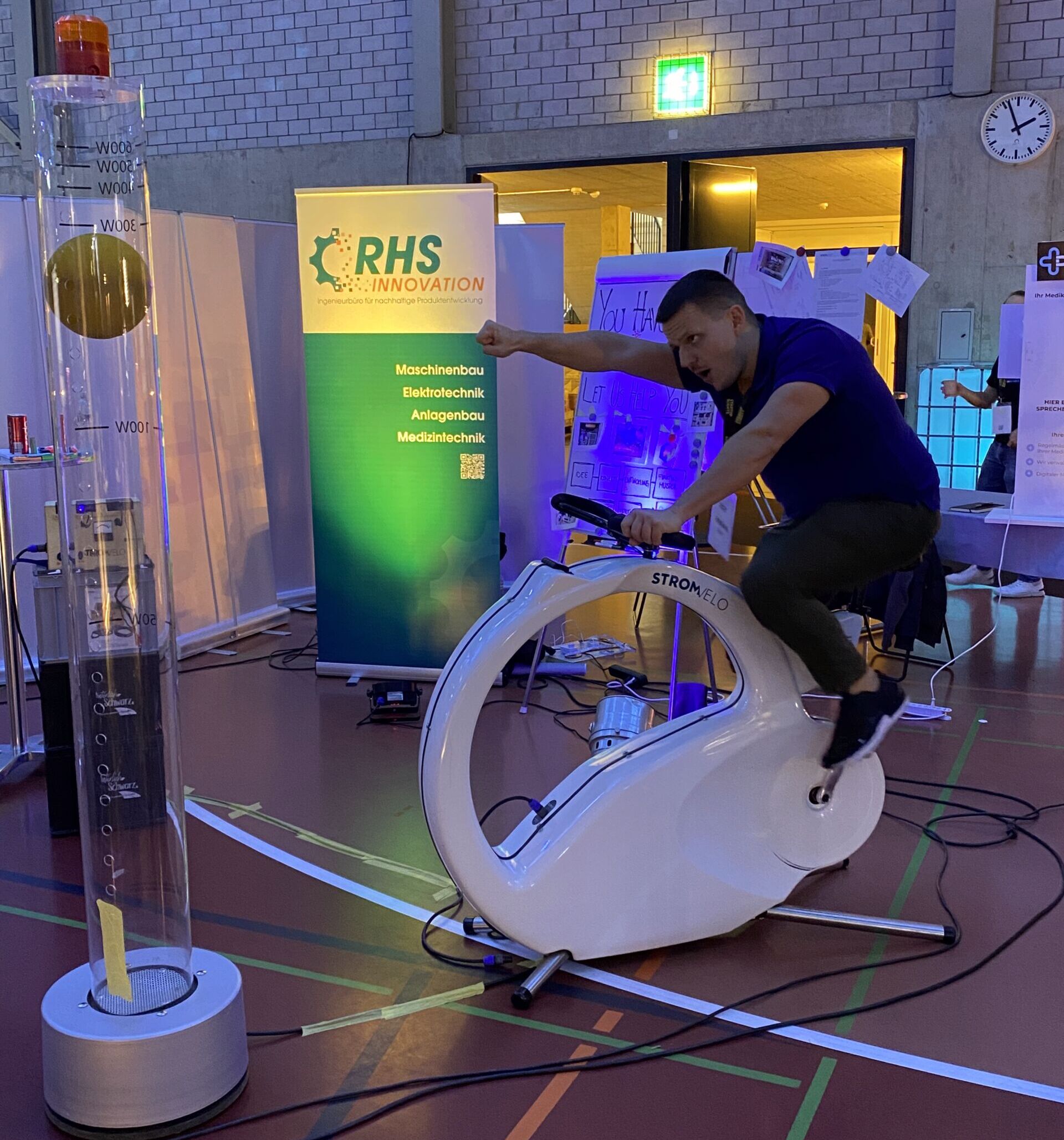 IMG_3454 | Strom-Velo Man rides a white exercise bike at an indoor expo with a green RHS Innovation banner nearby and a tall transparent cylinder on the left.