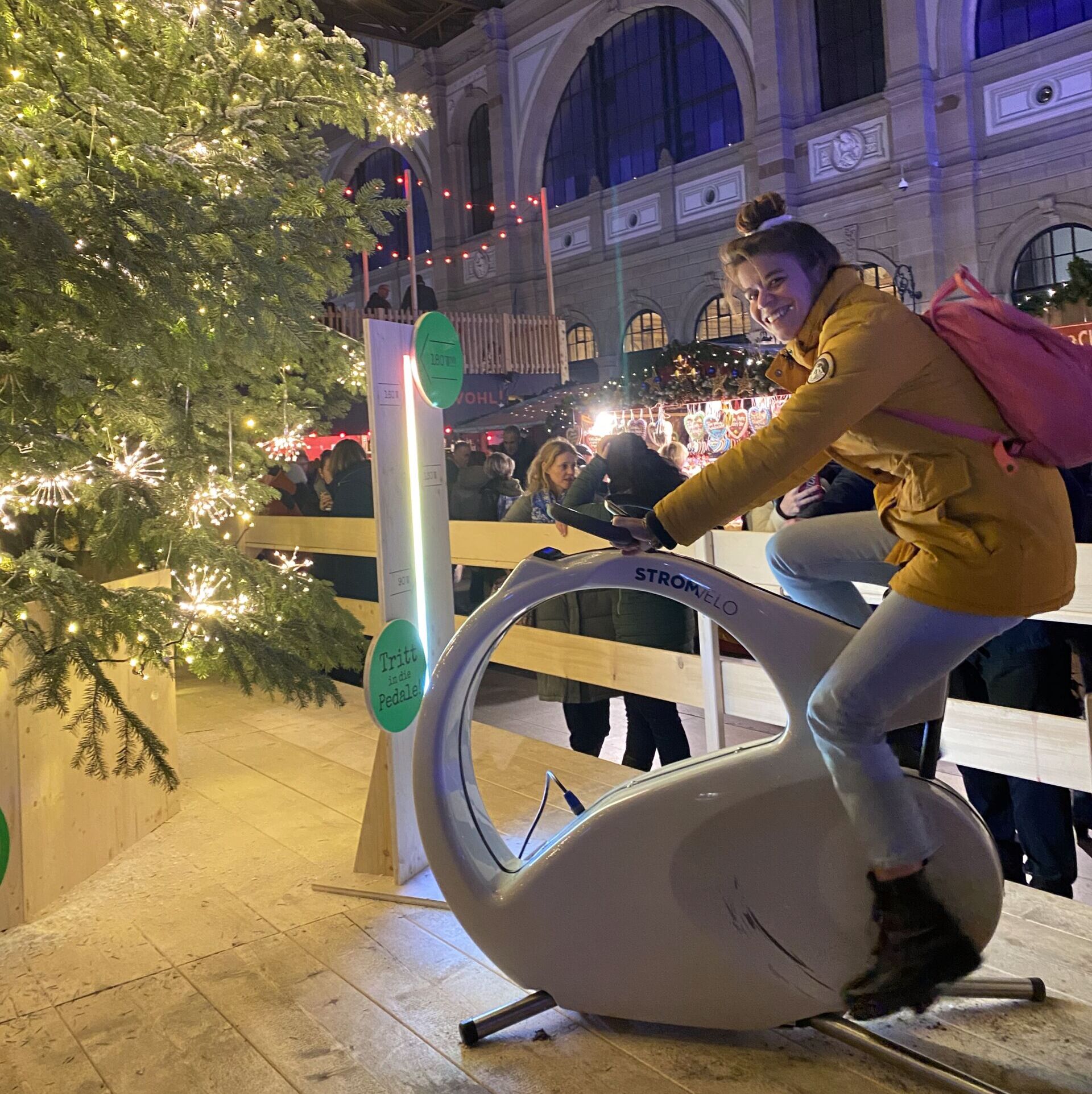 Woman in a yellow jacket rides a stationary bike at a Christmas market beside a lit, decorated tree.
