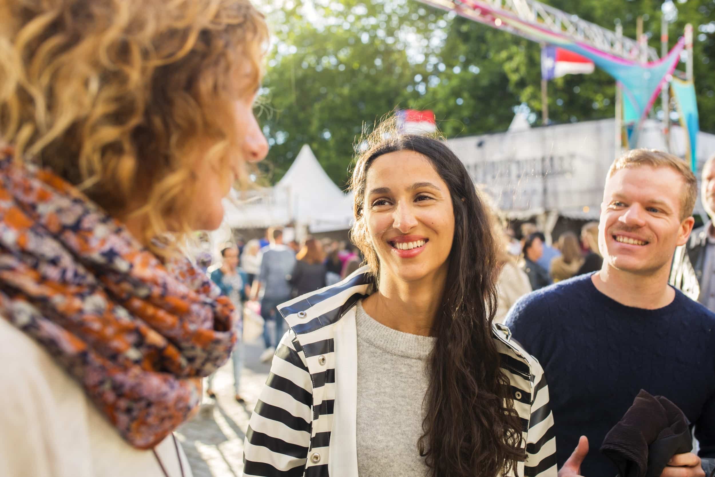 People smiling and chatting at an outdoor festival with colorful flags in the background.